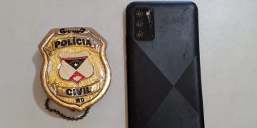 Celular preto ao lado de um distintivo da Polícia Civil de Rondônia.